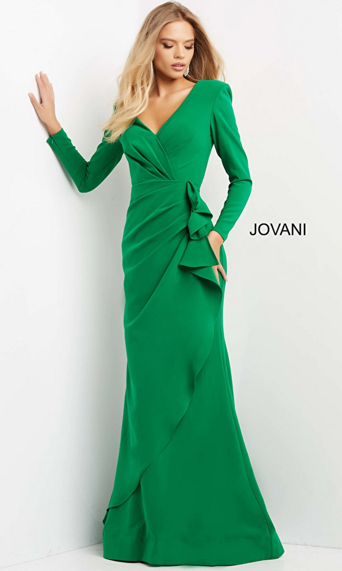 A woman stands against a white background wearing the Jovani 06995, a long-sleeve formal designer dress in emerald green with a deep V-neckline and side ruffle detail. The word
